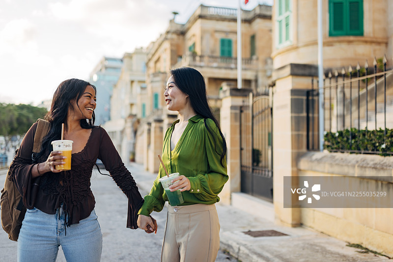 两位不同背景的挚友在城市街道上漫步，享受着欢乐时光。她们手牵着手，喝着健康的果昔，庆祝着彼此的友谊。图片素材