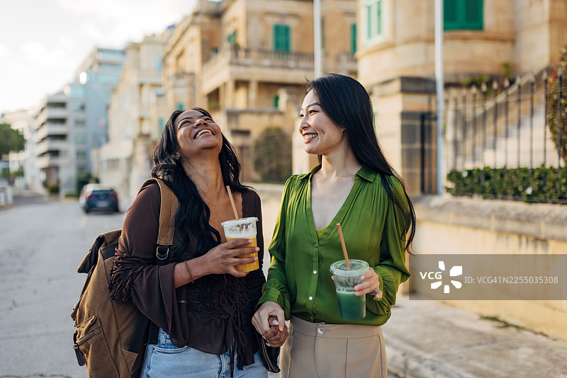 两位不同种族肤色的女性，在风景如画的城市区域手牵手、开怀大笑地走着，她们手里拿着最爱的外卖冰沙，享受着真实而美好的连接时刻。图片素材