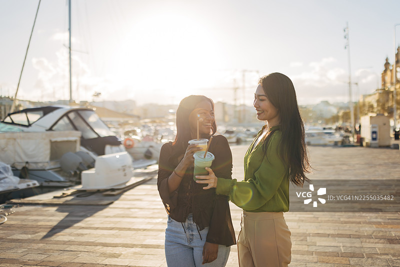 两位无忧无虑的女性，手持健康的即取饮品，享受着彼此的连接时刻。她们漫步在风景优美的石板铺就的水滨，代表着新一代注重健康和有意识的旅行者。图片素材