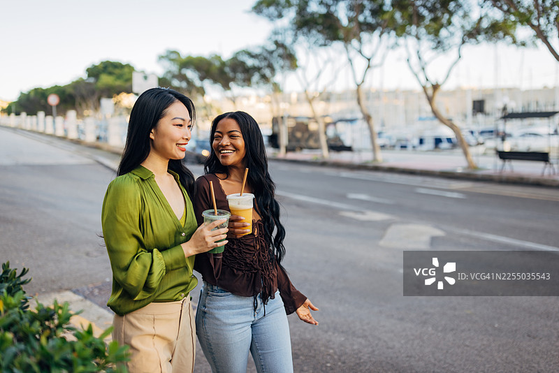 两位不同种族背景的女性，在一条迷人的城市街道上边走边享受阳光明媚的周末时光，她们手中拿着健康的鲜榨果汁。图片素材