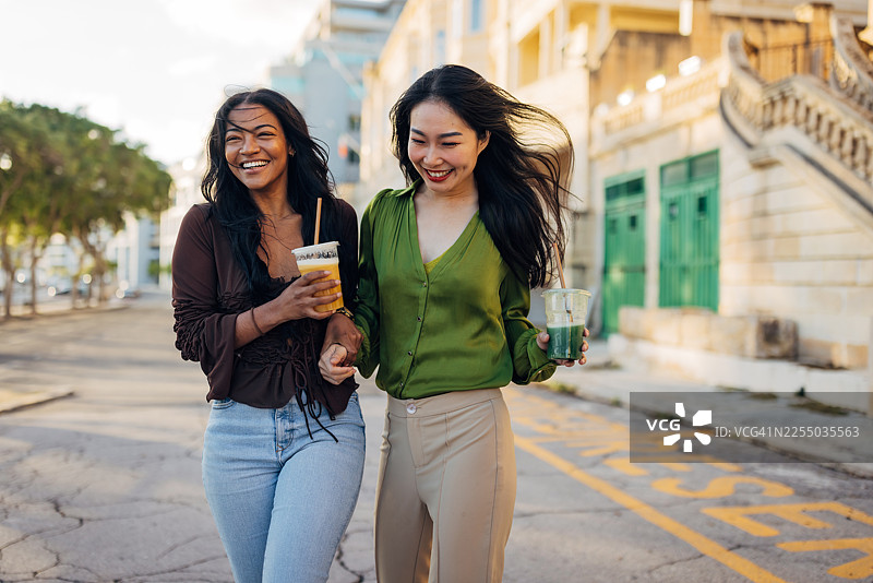 两位不同肤色的女性在城市中享受阳光明媚的一天。她们手持外带的绿色和橙色冰沙，在优雅的历史建筑前漫步，并开怀大笑。图片素材