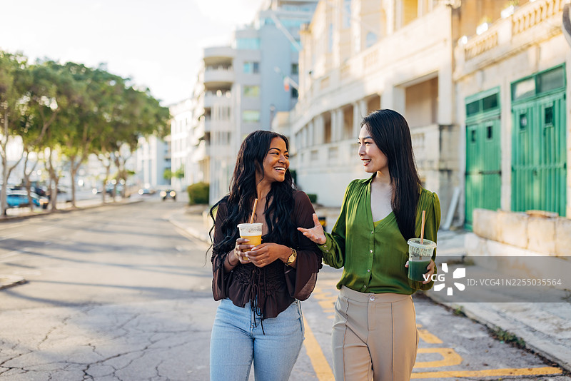 两位不同族裔的挚友在经典石砌建筑旁边走边愉快地交谈，他们一边喝着新鲜水果冰沙，展现出度假期间健康积极的生活方式。图片素材