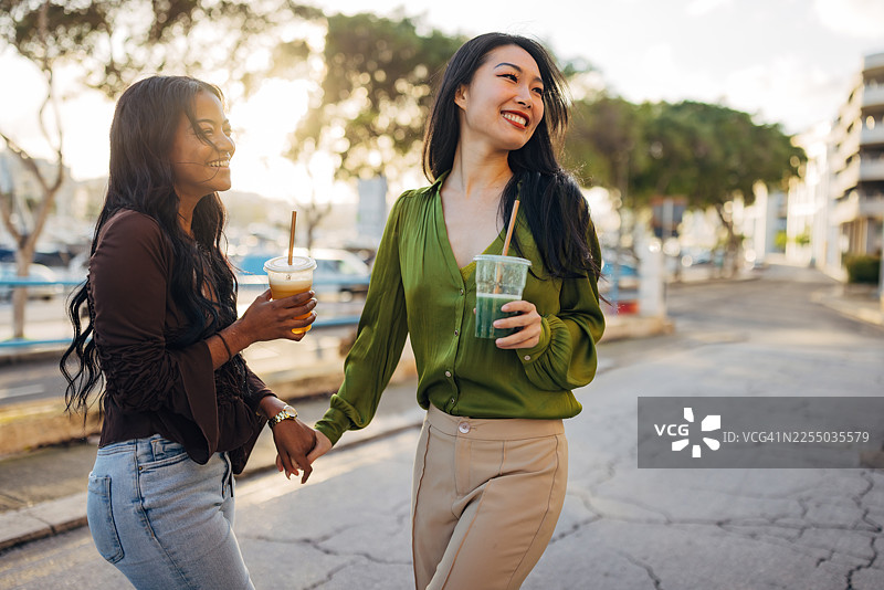 两位不同肤色的女性正在享受城市度假，她们走在阳光明媚的街道上，周围是历史建筑，手中拿着色彩缤纷的水果冰沙，享受着她们真实而欢乐的时刻。图片素材