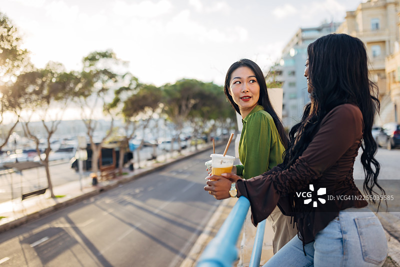 两位不同族裔的女性倚靠在蓝色栏杆上，面带喜悦的笑容互相凝视。她们在阳光明媚的城市里，手持外卖的绿色和橙色冰沙，背景是历史建筑和绿树成荫的街道。图片素材