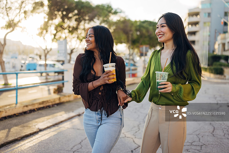 两位不同肤色的女性正在进行城市度假旅行，她们走在阳光明媚的街道上，周围是历史建筑，手里拿着色彩鲜艳的水果冰沙，享受着一段 candid 的连接与欢笑的时刻。图片素材