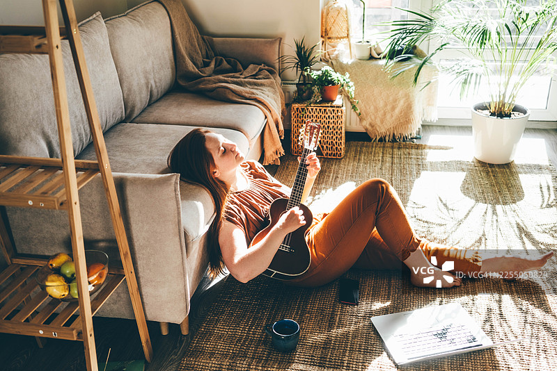 一位女士坐在阳光洒满的地毯上，闭着眼睛弹奏尤克里里。这个温馨的居家场景捕捉了在家中创意放松和宁静独处的片刻。图片素材