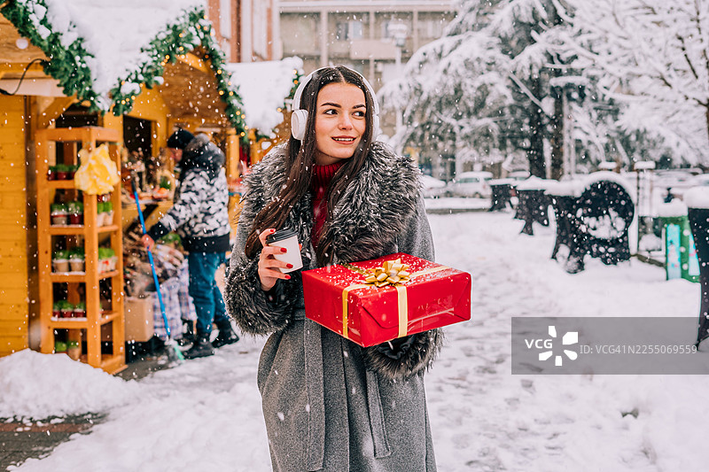 年轻女子在城市雪景圣诞集市享受节日气氛，手提礼物，品尝热巧克力。节日彩灯闪烁，身穿冬装，氛围温馨。图片素材