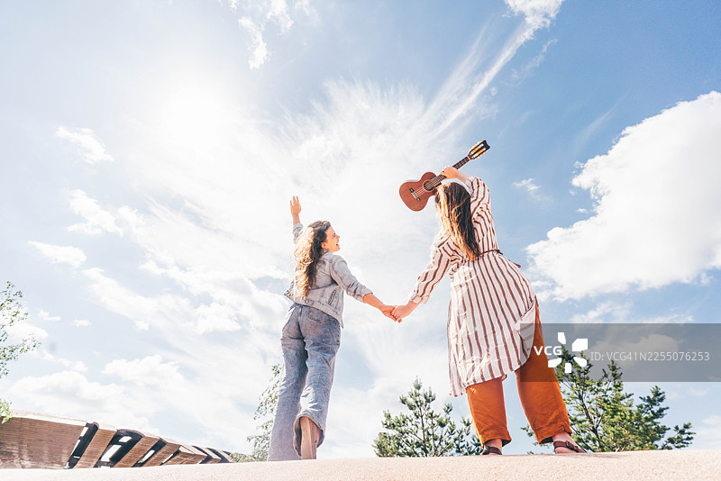 两位女性朋友手牵着手，在明媚的天空下，高高举起尤克里里，显得十分得意。图片素材
