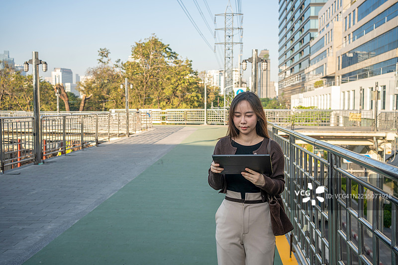 一位年轻的亚洲女商人自信地站在城市中，使用数字平板电脑，背景是高楼大厦和摩天大楼。图片素材