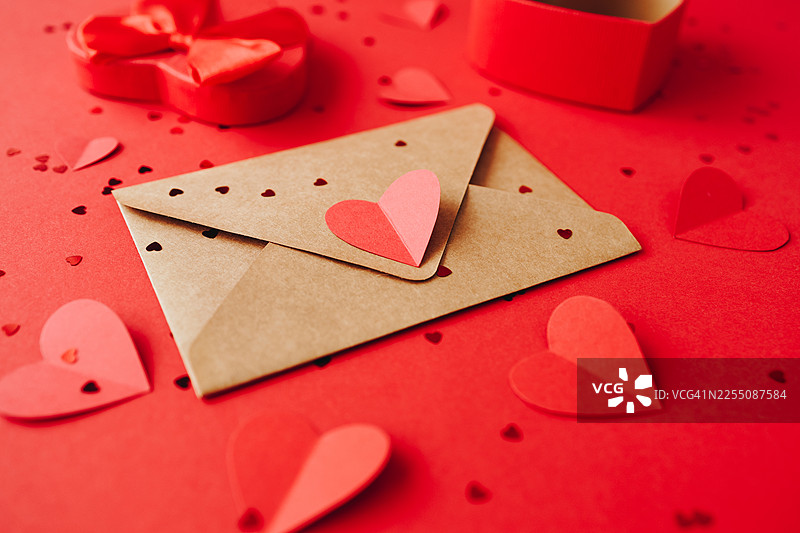 红底上带有纸质爱心的棕色牛皮纸信封，周围散落着彩纸和礼盒。图片素材