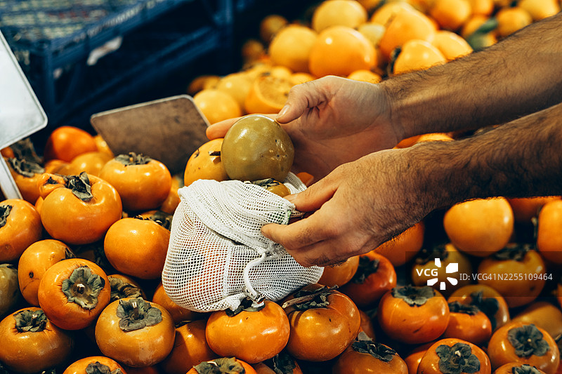 男士手中从堆里挑选成熟的柿子，并放入环保网袋中。零浪费。图片素材