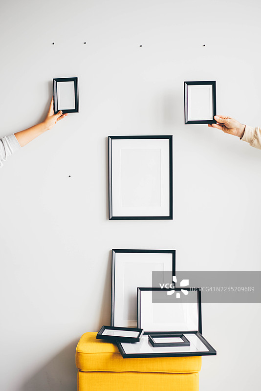 一个人正在纯白色背景的墙上摆放不同尺寸的黑色画框，布置成一组照片墙。图片素材