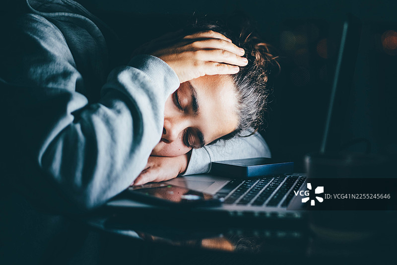 夜幕降临，一位疲惫的女子趴在办公桌上睡着了，旁边是发光的笔记本电脑，身后是黑暗的窗户。图片素材