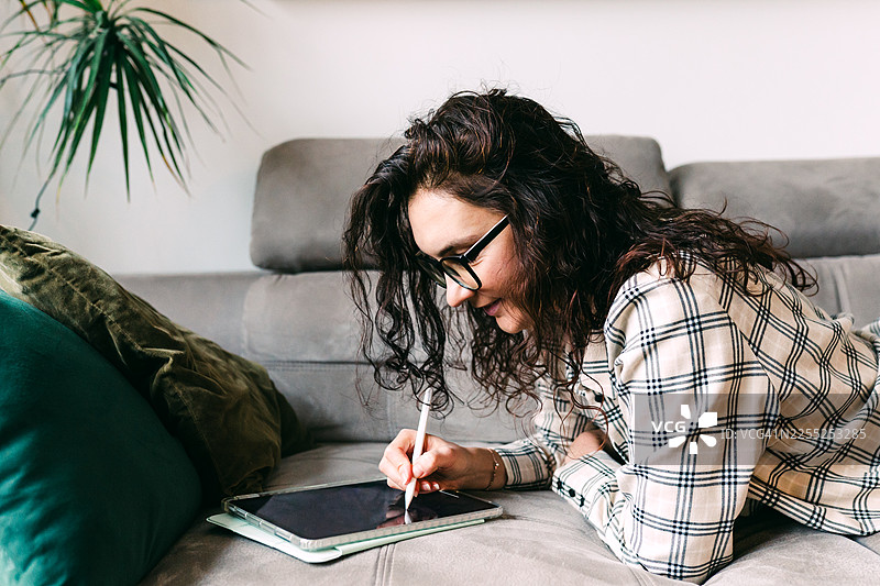 女子在家舒适地躺在沙发上，使用触控笔操作数码平板电脑图片素材