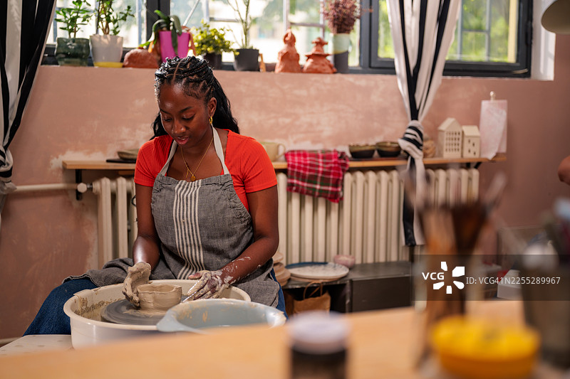 年轻女子在陶轮上熟练地揉捏陶土，在她充满活力和启发的车间里创作一件独特的作品，周围摆满了工具、材料和已完成的作品。图片素材