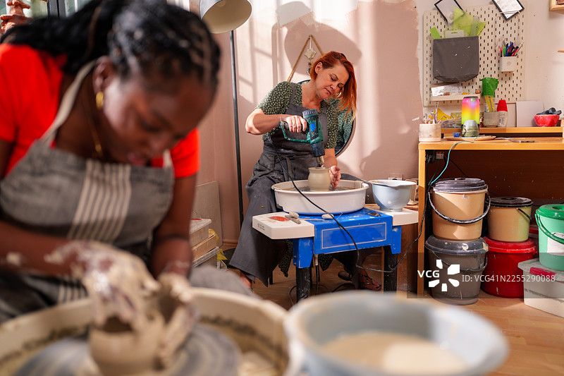 一位黑人女性和一位红发女性在明亮的工坊里使用陶轮制作陶土，她们使用工具，周围摆放着装有材料的桶。图片素材