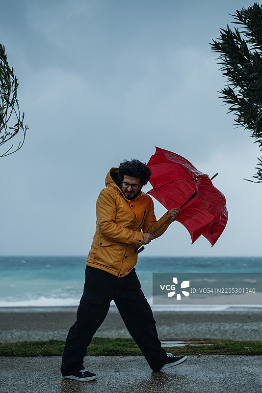 一名身穿黄色夹克的男子在海边，于风暴中挣扎着撑开一把红伞图片素材