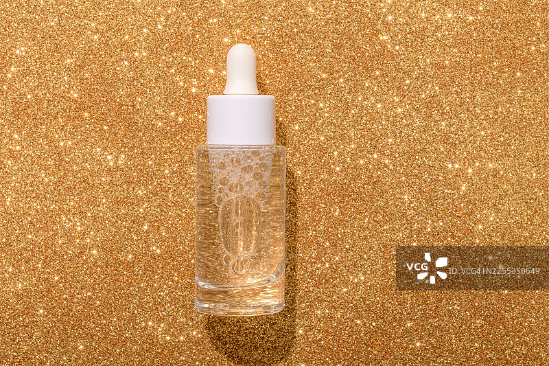 一个透明的滴管瓶放置在闪耀的金色闪光表面上，传达出奢华和高端护肤的质感。非常适合化妆品、精华液和美容品牌，营造出迷人、节日般的氛围。图片素材