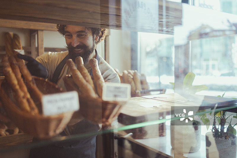 一位穿着围裙和手套的快乐面包师，在一家乡村风格的手工烘焙坊里，将刚出炉的面包放入柳条篮中。图片素材