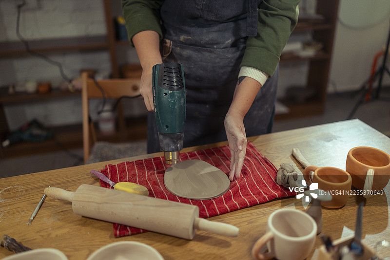 陶艺工作室里，一位女性陶艺师正在使用热风枪处理粘土，其身份无法辨认图片素材