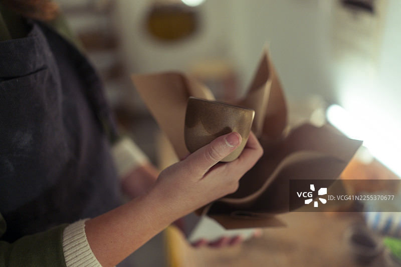 一位模糊了面容的女性企业家兼陶瓷艺术家正小心翼翼地将一个烧制完成、施了釉的陶杯放入一个装有牛皮纸的礼品盒中图片素材