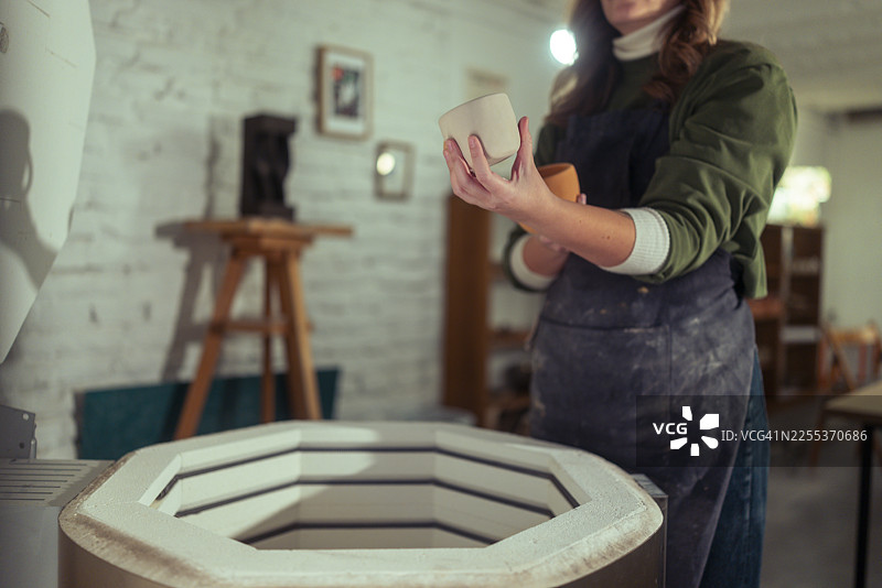 一位穿着围裙、无法辨认性别的陶瓷艺术家，手持两个手工陶艺马克杯，准备送入窑炉烧制图片素材