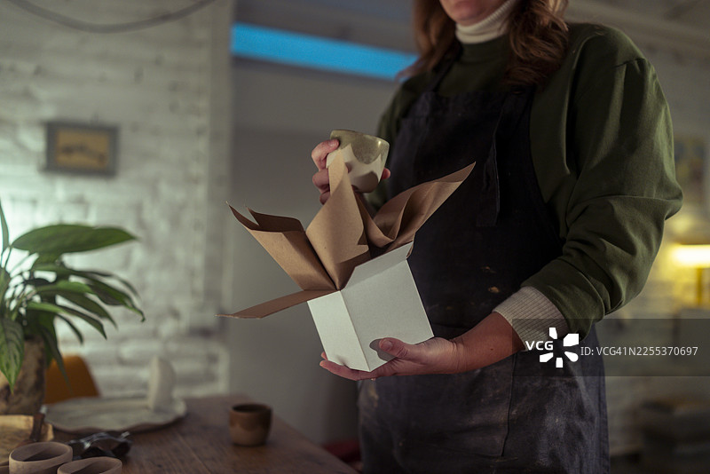 一位看不清面容的女性陶瓷艺术家正小心翼翼地将一个已烧制上釉的陶杯放入装有牛皮纸的礼品盒中图片素材