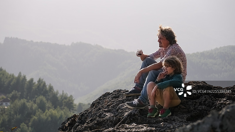 父亲和十几岁的孩子在山脊上，一边欣赏风景一边分享饮品图片素材
