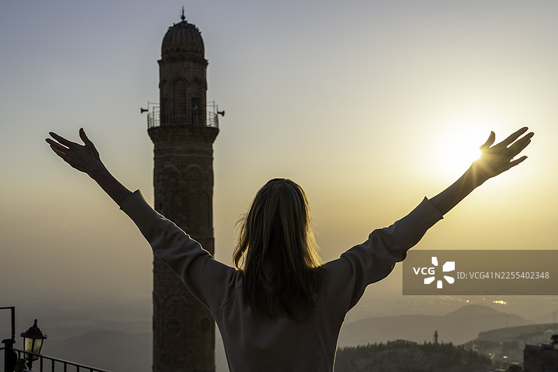 土耳其马尔丁日落时女子举起双臂，背景是历史悠久的宣礼塔图片素材