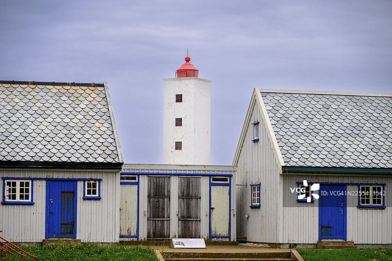 在灰色的天空下,一座灯塔矗立在两栋蓝白相间的房屋之间,这里是挪威芬马克郡贝勒福格乡下的Kjølnes灯塔,有小窗户和栏杆。图片素材