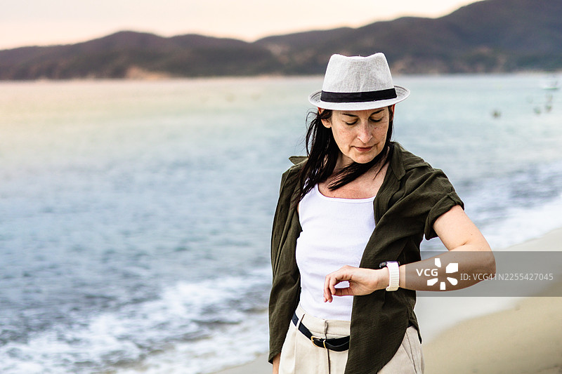戴着草帽的女性在海边，手臂上的连续血糖监测仪、智能手表、手机和防水CGM保护让她更轻松地旅行和管理糖尿病。图片素材
