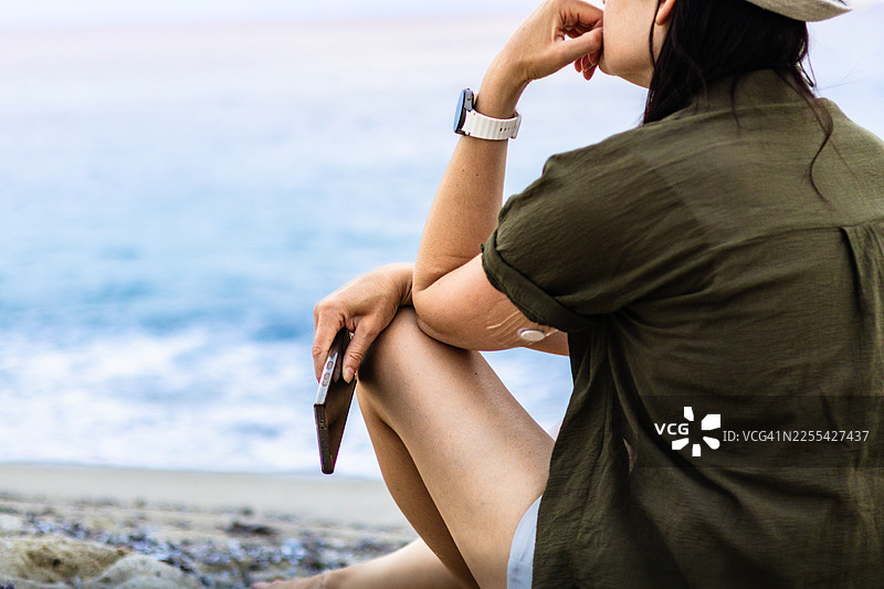 戴着草帽的女性在海边，她手臂上的连续血糖监测仪、智能手表、手机以及防水CGM保护装置，让糖尿病患者的旅行和生活更加轻松。图片素材