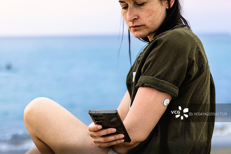 戴着草帽的女性在海边，手腕上的连续血糖监测仪（CGM）、智能手表、手机以及防水CGM保护套让她能够更轻松地旅行和管理糖尿病。图片素材