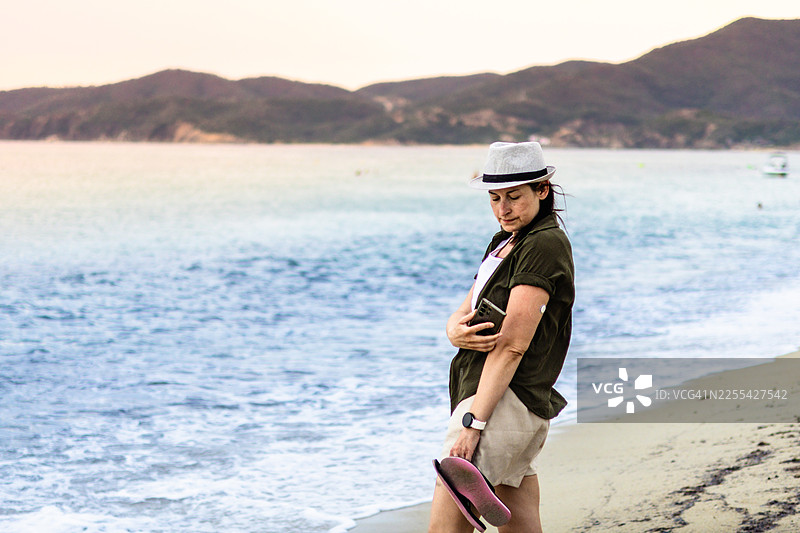 戴着草帽的女士在海边。有了手臂上的持续葡萄糖监测仪、智能手表、手机和防水CGM保护套，糖尿病患者的旅行和生活都变得更轻松。图片素材