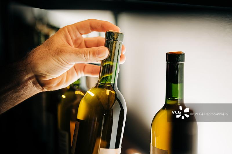 特写镜头，一只手正从极简的背光酒架上小心翼翼地挑选一瓶白葡萄酒。温暖的灯光和人手的触碰，营造出专业和精心挑选的氛围。图片素材