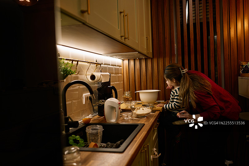 温馨的厨房里，一位母亲和孩子在温暖的灯光下一起烘焙饼干图片素材