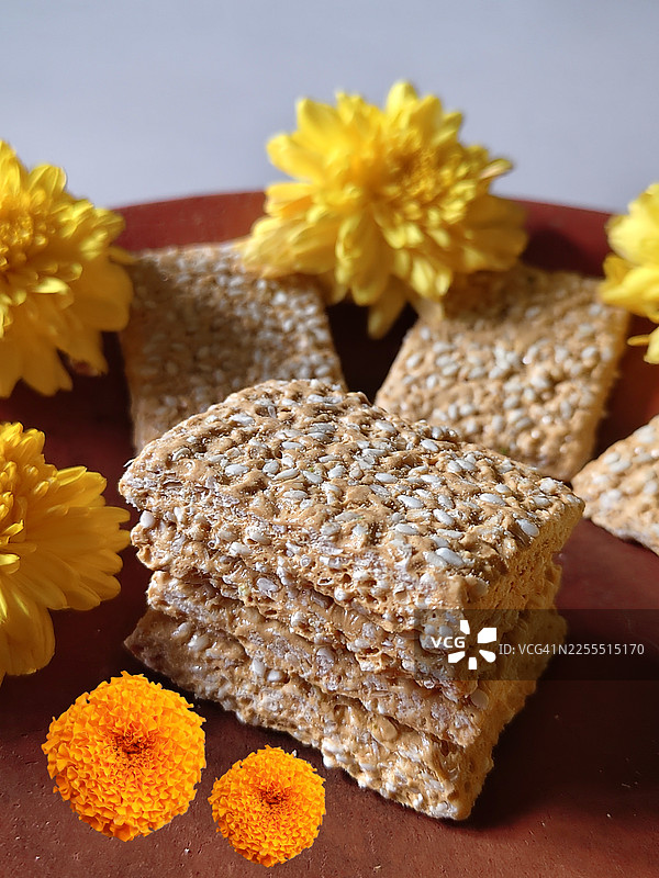 泰尔奇奇、提尔古尔、加扎克（一种由芝麻和粗糖/糖制成的传统印度干甜点）的摄影作品，装饰性地摆放在陶盘上，周围环绕着黄色的花朵——摩卡拉·桑克兰蒂节图片素材