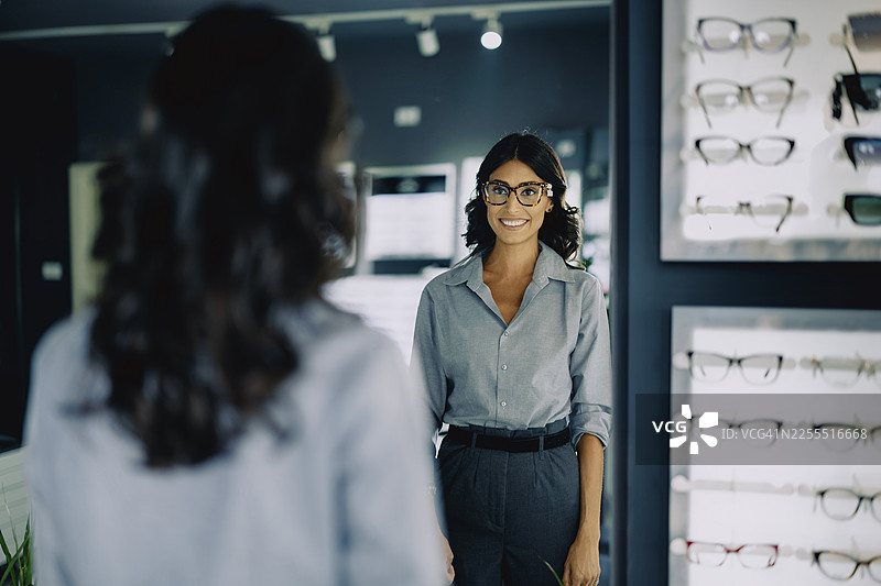 一家现代眼镜店里，一位迷人的年轻女子正在试戴眼镜。她站在陈列架旁，认真地挑选着不同的镜框。店内明亮而时尚，背景中可以看到各种各样的眼镜。图片素材