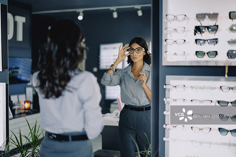 一位迷人的年轻女子在现代眼镜店试戴眼镜。她站在展示架旁，若有所思地挑选着不同的镜框。店内明亮时尚，背景中可以看到各种各样的眼镜。图片素材