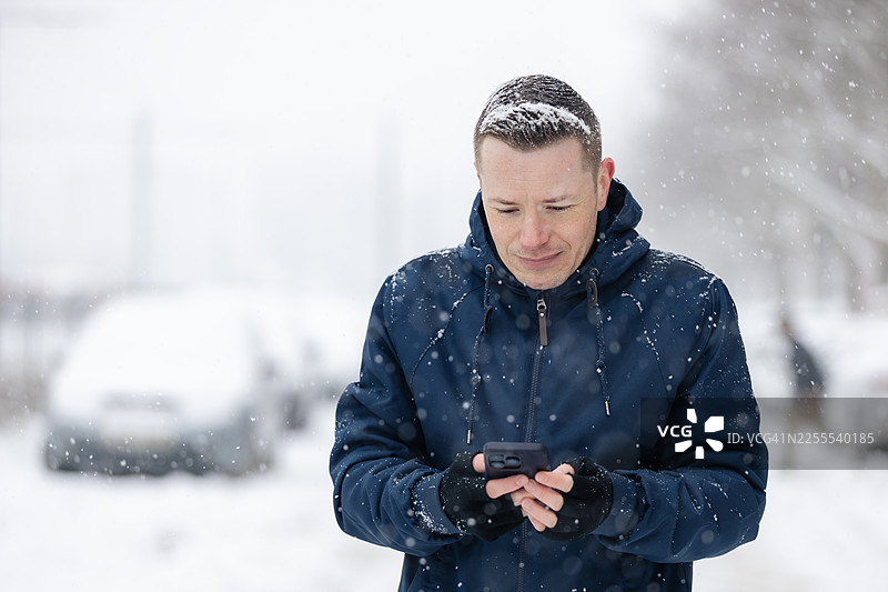 在寒冷的冬日，一位男士在雪中的城市里边走边看手机图片素材