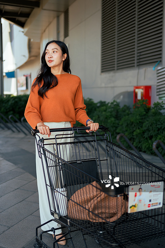 年轻的亚洲女性在超市外推着空的购物车，望向别处。着重于消费主义和城市活动图片素材