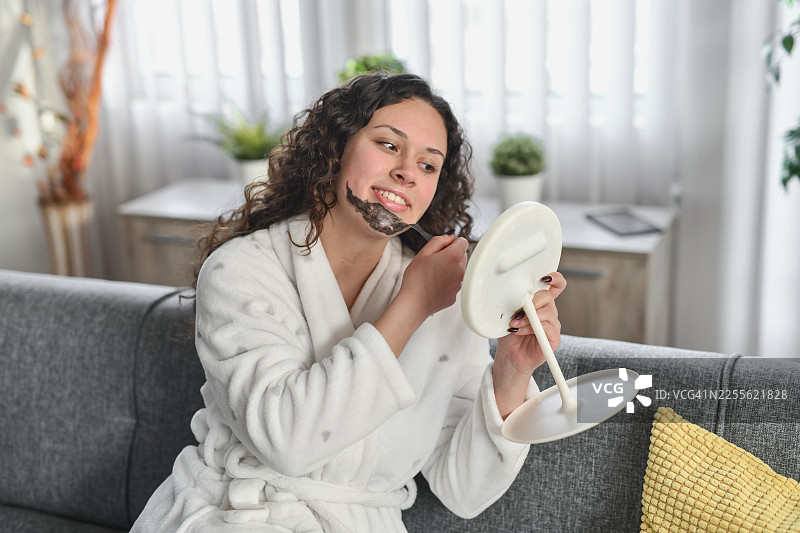 身穿白色浴袍的女性在家享受水疗日，旁边有镜子和舒适的沙发图片素材