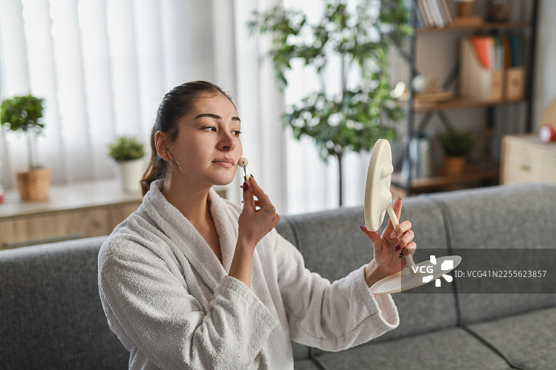 身着浴袍的女性在家中享受宁静的居家水疗日，使用玉石滚轮美容仪图片素材