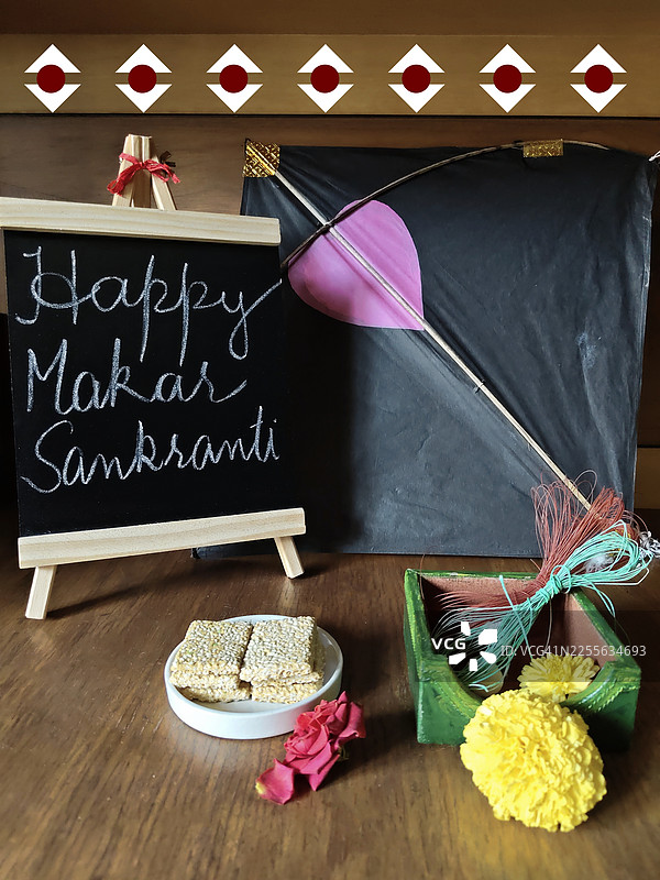 庆祝马卡尔·桑克兰蒂/北方节的物品照片，木制画架上的黑色小黑板写着“马卡尔·桑克兰蒂快乐”，万寿菊、玫瑰花、Gajak（一种印度甜点）、芝麻脆饼、印度脆饼、风筝。图片素材