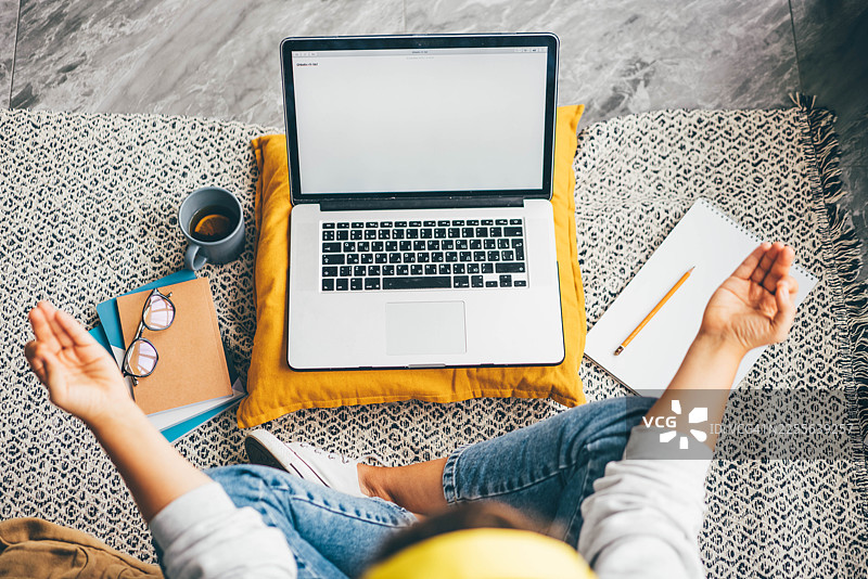 一位女性盘腿坐在有图案的地毯上，双手张开，呈冥想姿势，对着放在黄色坐垫上的笔记本电脑。图片素材