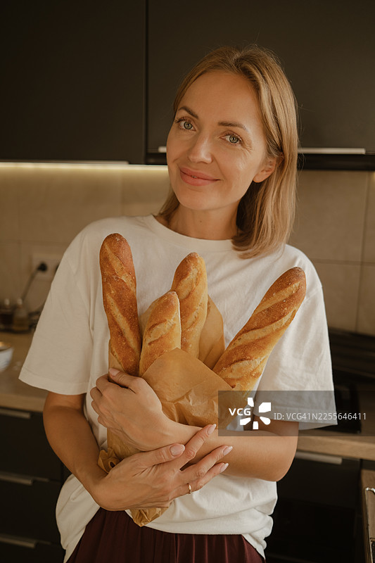 一位女士在家烘焙面包，她手中拿着法棍面包，脸上带着微笑。图片素材