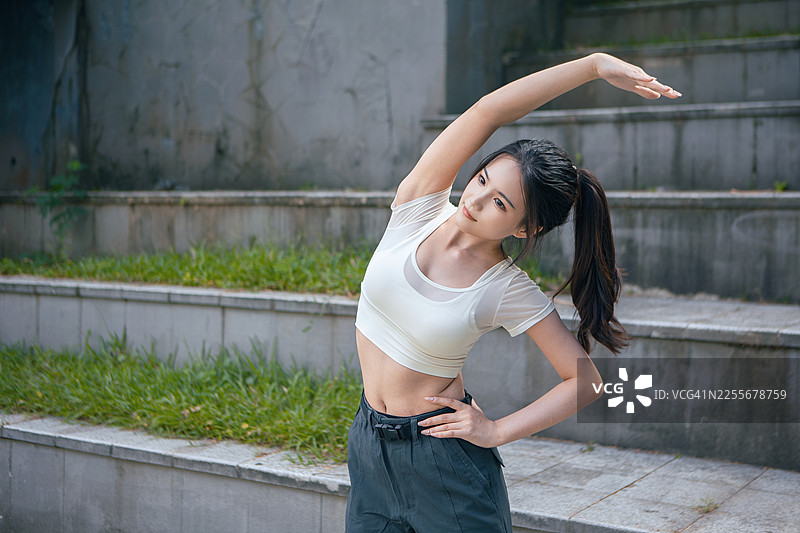 亚洲年轻女子在公园户外健身时伸展手臂，身穿运动背心，积极训练，展现健康生活方式、锻炼习惯和养生理念。图片素材