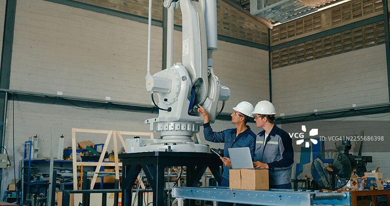 两位年轻的工程师在工厂操作大型机械臂，使用笔记本电脑和平板电脑管理机器。机器人工业工厂。图片素材