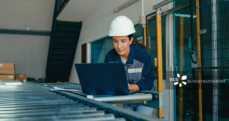 年轻的女工程师戴着安全帽，在工厂使用笔记本电脑分析数据并管理运营。机器人工业工厂。图片素材