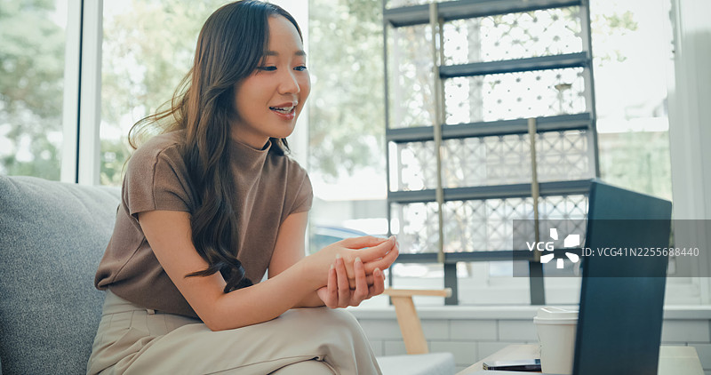 一位戴着耳机、身穿居家服的年轻亚洲女性，在舒适的咖啡馆里通过笔记本电脑进行视频通话，她一边挥手一边享用咖啡。咖啡馆生活方式。图片素材